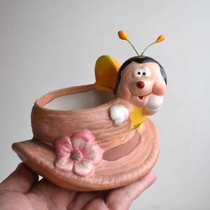 Könnte beinhalten: Ein Keramik-Pflanzgefäß in Form eines breitkrempigen Hutes mit einer rosa Blume auf der Vorderseite. Eine Biene im Cartoon-Stil mit schwarzem Kopf, gelben Fühlern und weißen Händen ruht auf dem Rand des Hutes. Der Hut ist hellbraun.