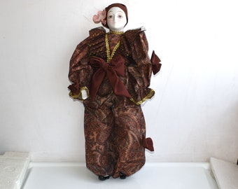 May include: A porcelain doll dressed in a brown and gold paisley patterned outfit with a large brown bow. The doll has a white face with painted features and a pink flower on its head.