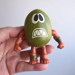 May include: A green egg-shaped toy with white eyes and a white, jagged mouth. The toy has orange arms and legs, with green bands around the wrists and ankles. The toy is held in a hand.