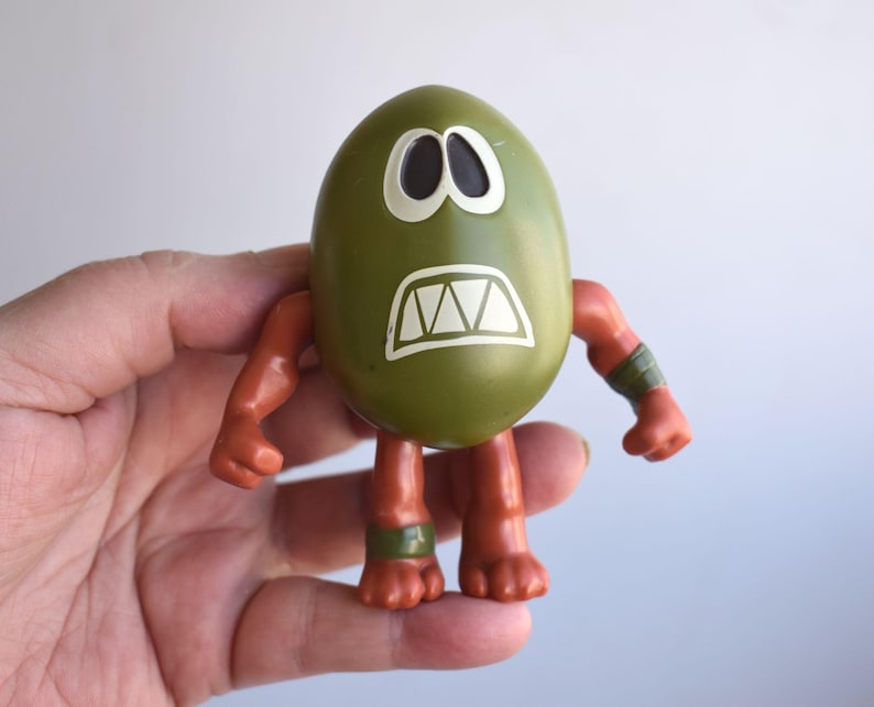 May include: A green egg-shaped toy with white eyes and a white, jagged mouth. The toy has orange arms and legs, with green bands around the wrists and ankles. The toy is held in a hand.