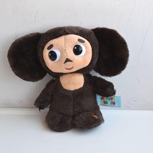 May include: A plush toy of a cartoon character with large, round eyes and oversized ears. The toy is primarily brown with a tan face and chest. A small blue tag is attached to the toy.