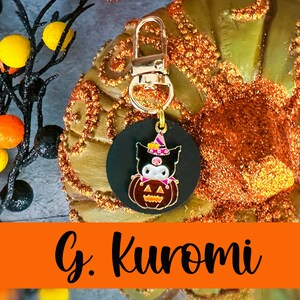 Pode incluir: Um chaveiro com tema de Halloween com um pingente circular preto com um personagem de desenho animado com chap&eacute;u de bruxa e uma ab&oacute;bora. O pingente &eacute; preso a um fecho dourado. O texto "G. Kuromi" &eacute; exibido em uma faixa laranja.