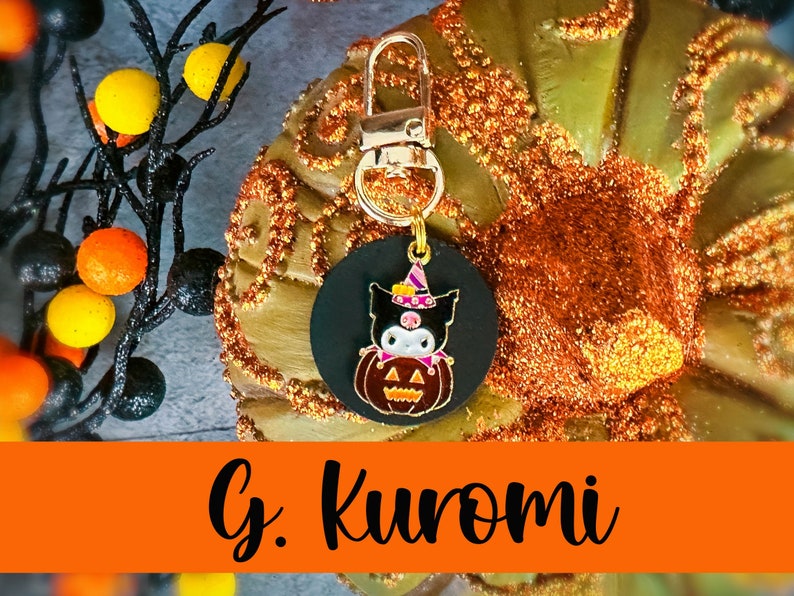 Pode incluir: Um chaveiro com tema de Halloween com um pingente circular preto com um personagem de desenho animado com chap&eacute;u de bruxa e uma ab&oacute;bora. O pingente &eacute; preso a um fecho dourado. O texto "G. Kuromi" &eacute; exibido em uma faixa laranja.