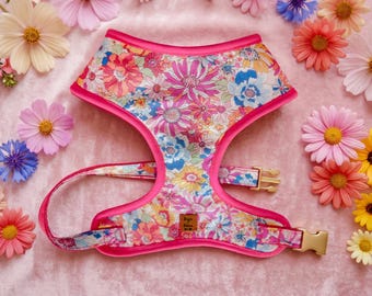 Puede incluir: Un arnés para mascota con un estampado floral vibrante de flores rosas, azules, amarillas y blancas. El arnés tiene bordes de color rosa intenso, hebillas de metal dorado y una pequeña etiqueta cuadrada marrón con el texto Juju Bear Co.
