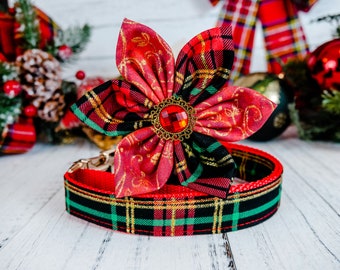 May include: A festive dog collar with a red, green, and black plaid pattern. The collar features a large, decorative flower with a red jewel center. The flower and collar are set against a holiday-themed background.