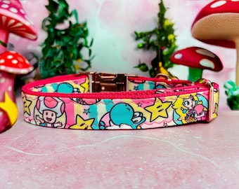 Op de afbeelding: Een roze hondenhalsband met een kleurrijk cartoonpatroon. De halsband heeft personages als Toad, Yoshi en Princess Peach, met gele sterren en een roségouden gesp. De halsband is ongeveer 2,5 cm breed.