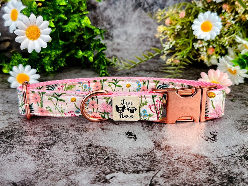 Puede incluir: Collar para perros con estampado floral sobre fondo rosa, adornado con flores rosas, azules y verdes. Hebilla de oro rosa y peque&ntilde;a etiqueta de madera con la inscripci&oacute;n "Juju Nana" e ilustraciones de perros.