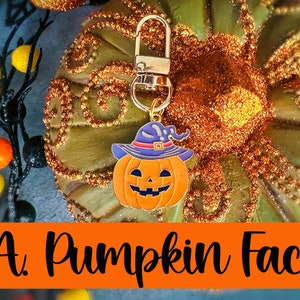 May include: A Halloween-themed charm featuring a smiling orange pumpkin face wearing a purple witch hat. The charm is attached to a gold-colored clasp. The background includes a decorative pumpkin and the text "A. Pumpkin Face".