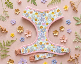 Puede incluir: Arnés para perros con estampado floral, con fondo azul claro y ribete rosa. El diseño presenta flores amarillas, azules y doradas. Una correa a juego está sujeta con una hebilla de oro rosa. Flores decorativas rodean el arnés.