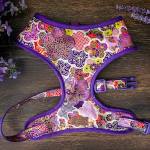 May include: Purple dog harness featuring a floral design in shades of pink, red, gold, and white. The harness has a buckle fastening and a D-ring for attaching a lead.