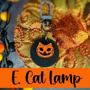 Pode incluir: Um chaveiro preto com fecho prateado e um pingente circular com um rosto de gato ab&oacute;bora laranja. O texto "E. Cat Lamp" &eacute; exibido em uma faixa laranja na parte inferior da imagem. O fundo inclui uma ab&oacute;bora dourada.