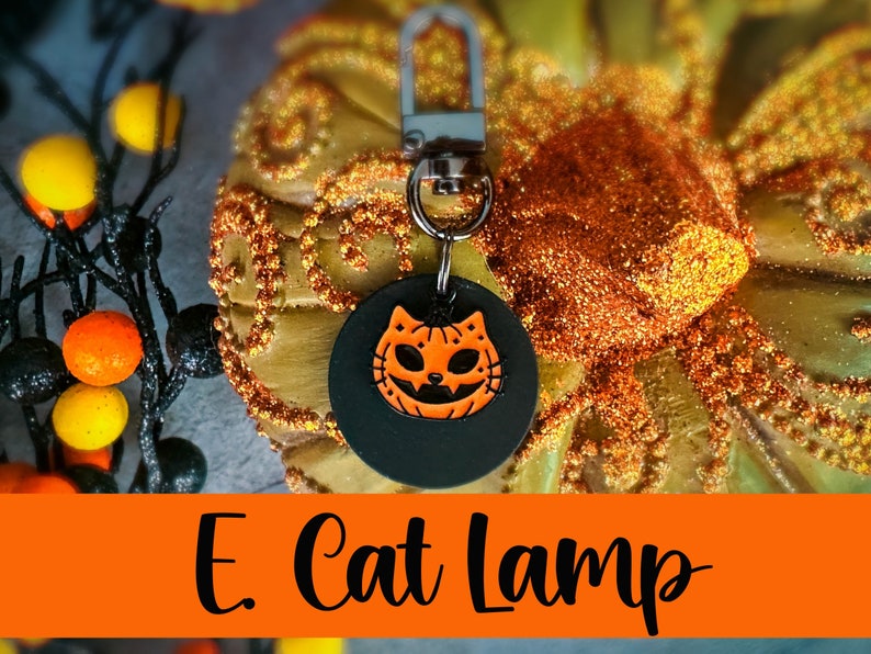 Pode incluir: Um chaveiro preto com fecho prateado e um pingente circular com um rosto de gato ab&oacute;bora laranja. O texto "E. Cat Lamp" &eacute; exibido em uma faixa laranja na parte inferior da imagem. O fundo inclui uma ab&oacute;bora dourada.