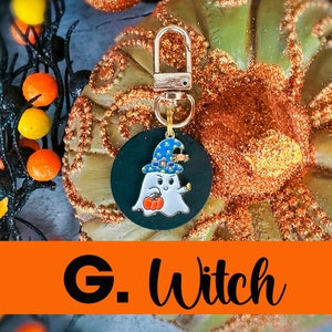 May include: A Halloween-themed keychain featuring a cartoon ghost wearing a witch hat and holding a pumpkin. The keychain has a gold-colored clasp and a black circular background. The text "G. Witch" is displayed on an orange banner.
