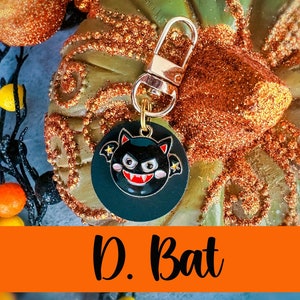May include: A round, black Halloween-themed charm with a gold clasp. The charm features a cartoon bat with a red mouth, white fangs, and star accents. The text "D. Bat" is displayed on an orange banner below the charm.