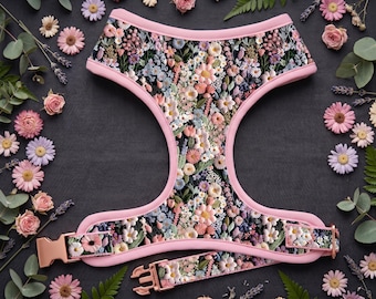 Puede incluir: Un arnés para perros y un juego de collar a juego con estampado floral. El arnés tiene un ribete rosa y un colorido estampado floral con una hebilla dorada. El conjunto está rodeado de flores y vegetación sobre un fondo oscuro.