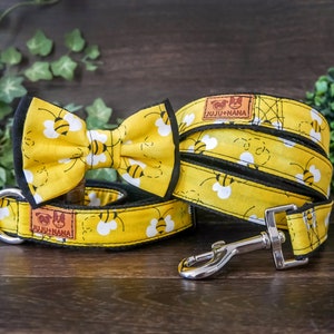 Pode incluir: Uma coleira para animal de estimação com laço a condizer e trela, todos com um padrão de abelhas a preto e branco. A coleira e a trela têm rebordos pretos, ferragens em metal prateado e uma etiqueta castanha com JUJU+NANA e dois ícones.