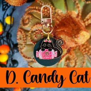 Pode incluir: Um pingente de gato preto com um design de embalagem de doces rosa, preso a um fecho dourado. O pingente &eacute; redondo e apresenta o texto "Halloween". As palavras "D. Candy Cat" est&atilde;o escritas em preto em uma faixa laranja.