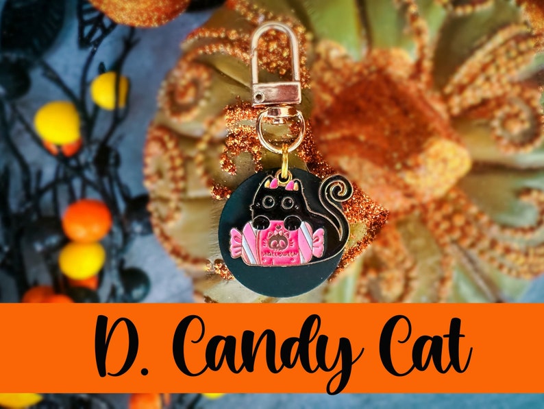 Pode incluir: Um pingente de gato preto com um design de embalagem de doces rosa, preso a um fecho dourado. O pingente &eacute; redondo e apresenta o texto "Halloween". As palavras "D. Candy Cat" est&atilde;o escritas em preto em uma faixa laranja.