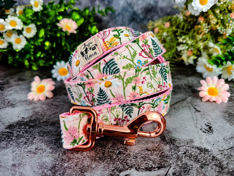 Puede incluir: Un juego de correa y collar para perros con un cierre de oro rosa. La correa y el collar presentan un fondo blanco con un estampado de flores rosas, amarillas y azules y hojas verdes. La marca "Juju Nana" est&aacute; en el collar.