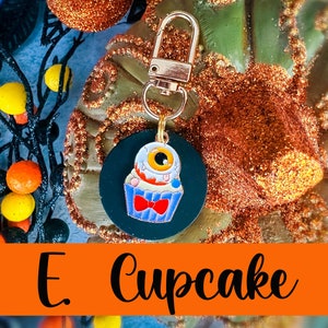 May include: A Halloween-themed keychain featuring a cupcake with an eyeball design. The cupcake has a blue wrapper, red bow tie, and a white and yellow eyeball. The keychain has a gold-colored clasp and a dark green backing. The word "Cupcake" is written in black.