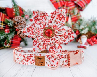 Puede incluir: Un collar blanco para perros y un accesorio floral a juego con un estampado de bayas rojas. El collar tiene herrajes de oro rosa y una etiqueta de cuero marrón que dice "Juju Nara". La flor tiene un centro de joya roja.