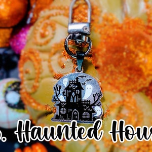 Pode incluir: Um chaveiro prateado com tema de Halloween. O pingente apresenta uma casa assombrada preta com fantasmas brancos em um fundo prateado. O texto "B. Haunted House" est&aacute; abaixo.