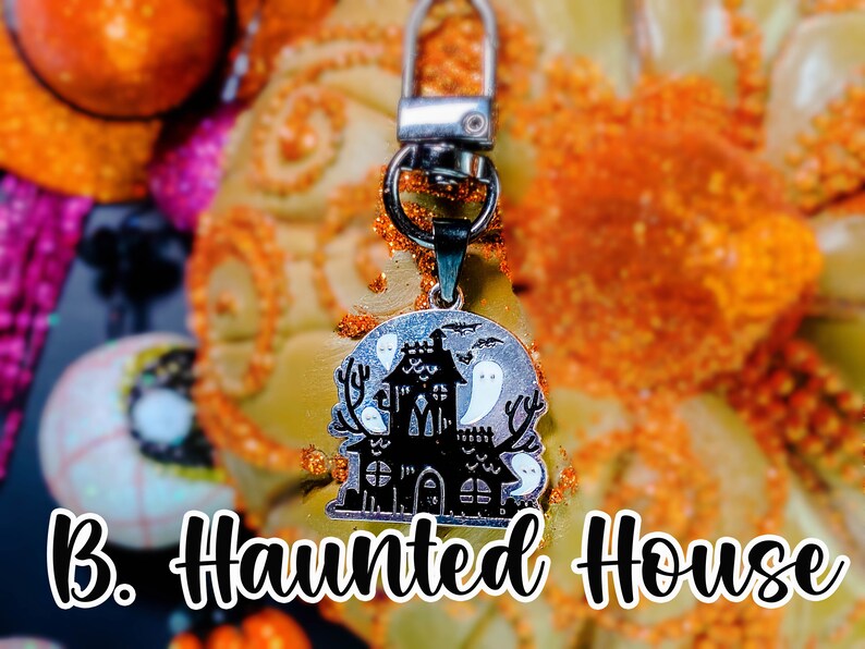 Pode incluir: Um chaveiro prateado com tema de Halloween. O pingente apresenta uma casa assombrada preta com fantasmas brancos em um fundo prateado. O texto "B. Haunted House" est&aacute; abaixo.