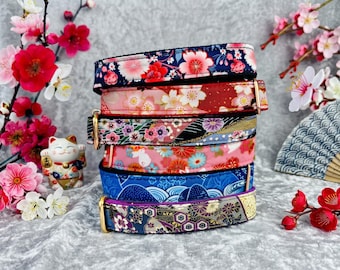 May include: A stack of six fabric pet collars featuring various Japanese-inspired patterns, including cherry blossoms, waves, and geometric designs. Each collar is equipped with a gold-toned metal buckle and hardware for secure fastening.