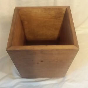 May include: A wooden box with a square shape and a light brown color. The box is empty and has a smooth surface.