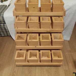 Puede incluir: Una estantería de madera con 15 macetas cuadradas de madera. La estantería está hecha de madera natural y tiene cuatro niveles. Las macetas son todas del mismo tamaño y están dispuestas en forma escalonada.