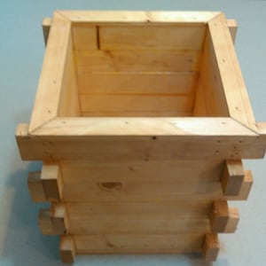 May include: A wooden box with a square shape and a light brown color. The box is made of several pieces of wood that are joined together. The box is empty and has a smooth surface.