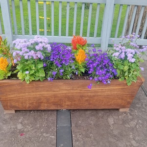 May include: A wooden planter box filled with colorful flowers. The flowers include vibrant orange, yellow, purple, and lavender blooms. The planter box is made of stained wood and sits on a gray surface.