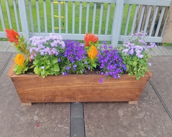 Op de afbeelding: Een houten plantenbak gevuld met kleurrijke bloemen. De bloemen omvatten feloranje, gele, paarse en lavendelkleurige bloemen. De plantenbak is gemaakt van gebeitst hout en staat op een grijze ondergrond.