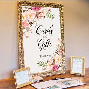 May include: A white sign with a gold frame and floral design. The sign reads "Cards and Gifts" with the text "Thank you" below.