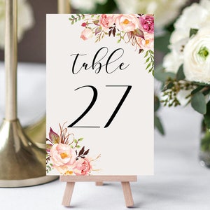 Może przedstawiać: Karta z numerem stołu 27 na małym drewnianym sztaludze. Karta zawiera słowo "table" w eleganckim skrypcie nad numerem 27. Kwiatowe akcenty akwarelowe w kolorze różowym, bordowym i zielonym zdobią górne i dolne rogi.