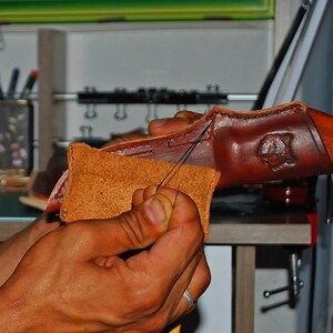 May include: Close-up of hands working on a brown leather product. A needle and thread are used to stitch together two pieces of leather. The leather has a natural, textured appearance, with a stamped design visible on the larger piece.