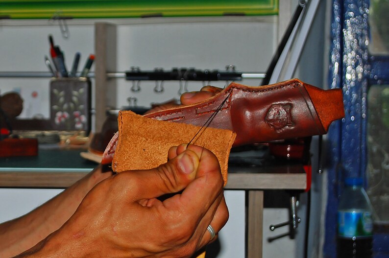 May include: Close-up of hands working on a brown leather product. A needle and thread are used to stitch together two pieces of leather. The leather has a natural, textured appearance, with a stamped design visible on the larger piece.