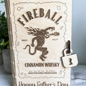 May include: Wooden box with a laser-etched design featuring the Fireball Cinnamon Whisky logo, a mythical creature, and the text "Happy Father's Day." Includes a wooden padlock closure.