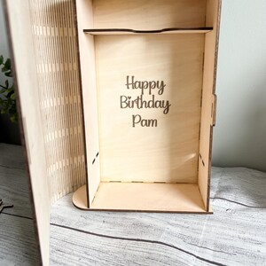 May include: A wooden box with an open lid, featuring the engraved text "Happy Birthday Pam" inside. The box is made of light-colored wood and has a shelf. The lid has vertical lines cut into it.