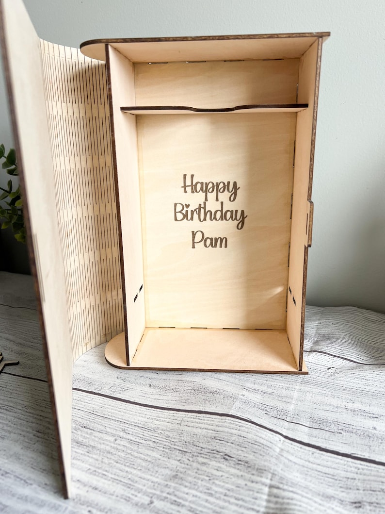 May include: A wooden box with an open lid, featuring the engraved text "Happy Birthday Pam" inside. The box is made of light-colored wood and has a shelf. The lid has vertical lines cut into it.