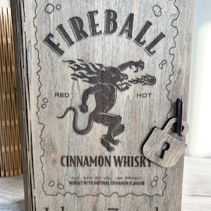May include: Wooden box with the word "FIREBALL" engraved on the front, along with a graphic of a mythical creature. The box also features the text "CINNAMON WHISKY" and "Johnny Zepeda". A small wooden padlock is attached.