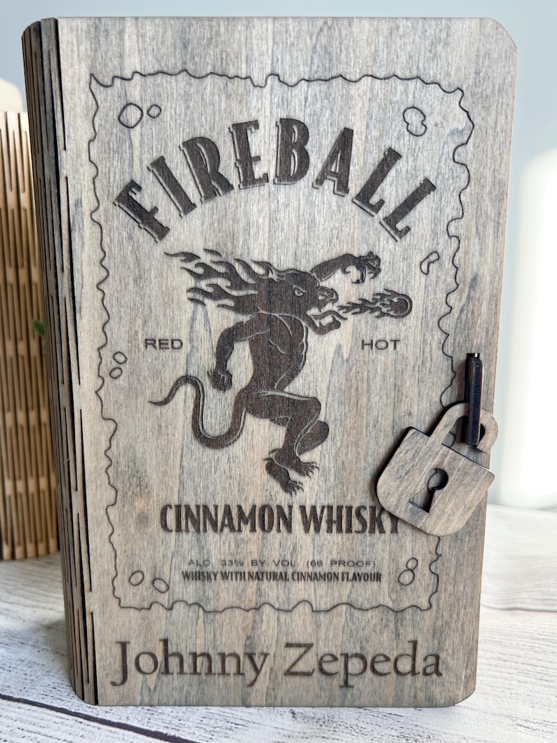May include: Wooden box with the word "FIREBALL" engraved on the front, along with a graphic of a mythical creature. The box also features the text "CINNAMON WHISKY" and "Johnny Zepeda". A small wooden padlock is attached.