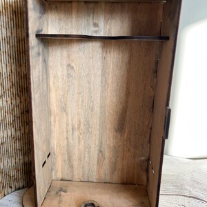 May include: A wooden box with a shelf and a decorative padlock. The box is made of light brown wood with visible grain. The padlock is also made of wood and is unlatched. The box has a rectangular shape.