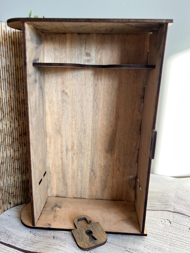 May include: A wooden box with a shelf and a decorative padlock. The box is made of light brown wood with visible grain. The padlock is also made of wood and is unlatched. The box has a rectangular shape.