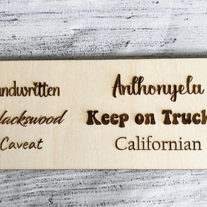 May include: A rectangular wooden sign with engraved text. The sign features the words "Handwritten", "Blackswood", "Caveat", "Anthonyela", "Keep on Truckin", and "Californian" in a dark brown color.