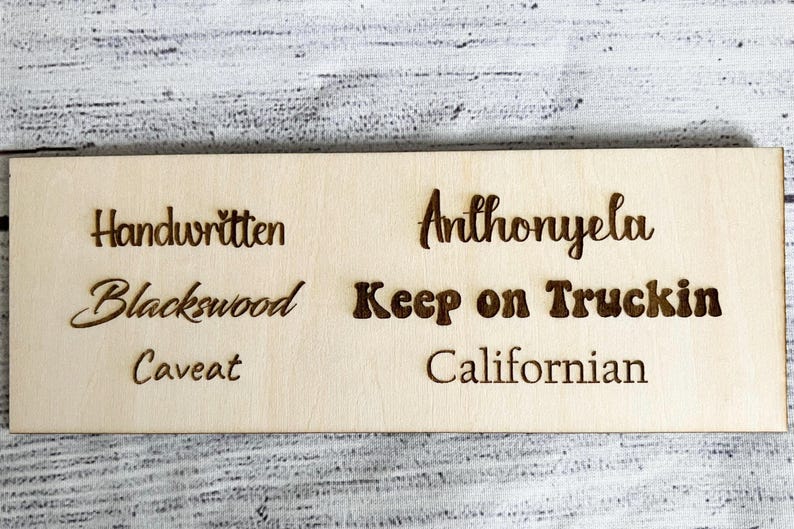 May include: A rectangular wooden sign with engraved text. The sign features the words "Handwritten", "Blackswood", "Caveat", "Anthonyela", "Keep on Truckin", and "Californian" in a dark brown color.