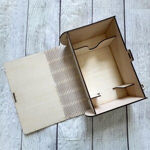 May include: A light-colored wooden box with an open lid, revealing interior compartments and shelves. The box is constructed from thin, laser-cut wood, with a latch closure. The background is a light-colored wooden surface.