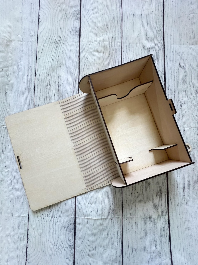 May include: A light-colored wooden box with an open lid, revealing interior compartments and shelves. The box is constructed from thin, laser-cut wood, with a latch closure. The background is a light-colored wooden surface.