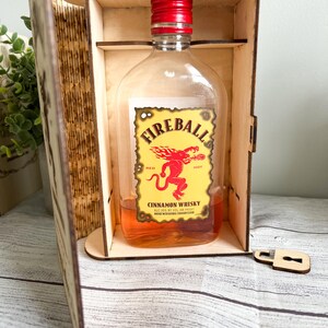 May include: A bottle of Fireball Cinnamon Whisky with a red cap and a yellow label featuring a red devil graphic. The bottle is displayed inside a wooden box with a lock and key.