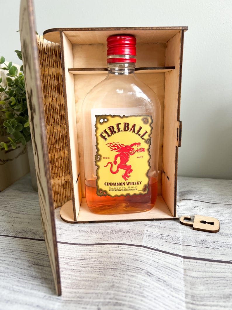 May include: A bottle of Fireball Cinnamon Whisky with a red cap and a yellow label featuring a red devil graphic. The bottle is displayed inside a wooden box with a lock and key.